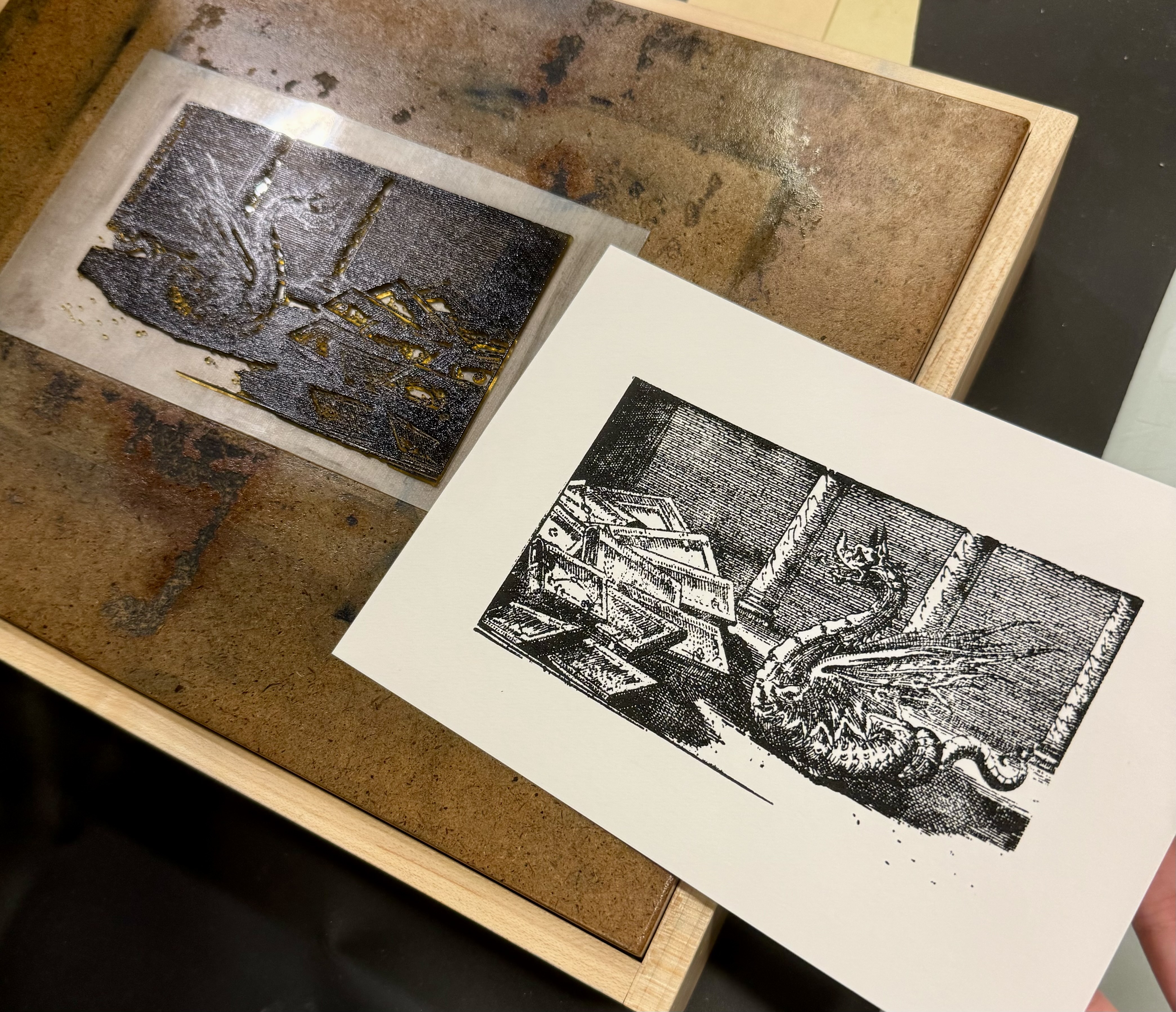 photograph of photopolymer plate inked and a print pulled on tabletop press. Plate depicts a dragon surrounded by books